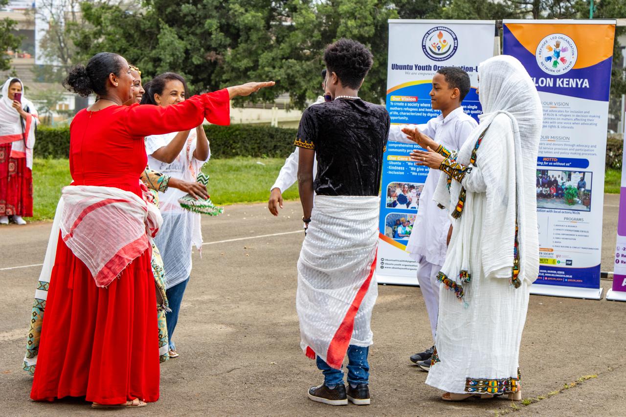 Community participants in red and white traditional clothing greeting one another near refugee-led organisation displays