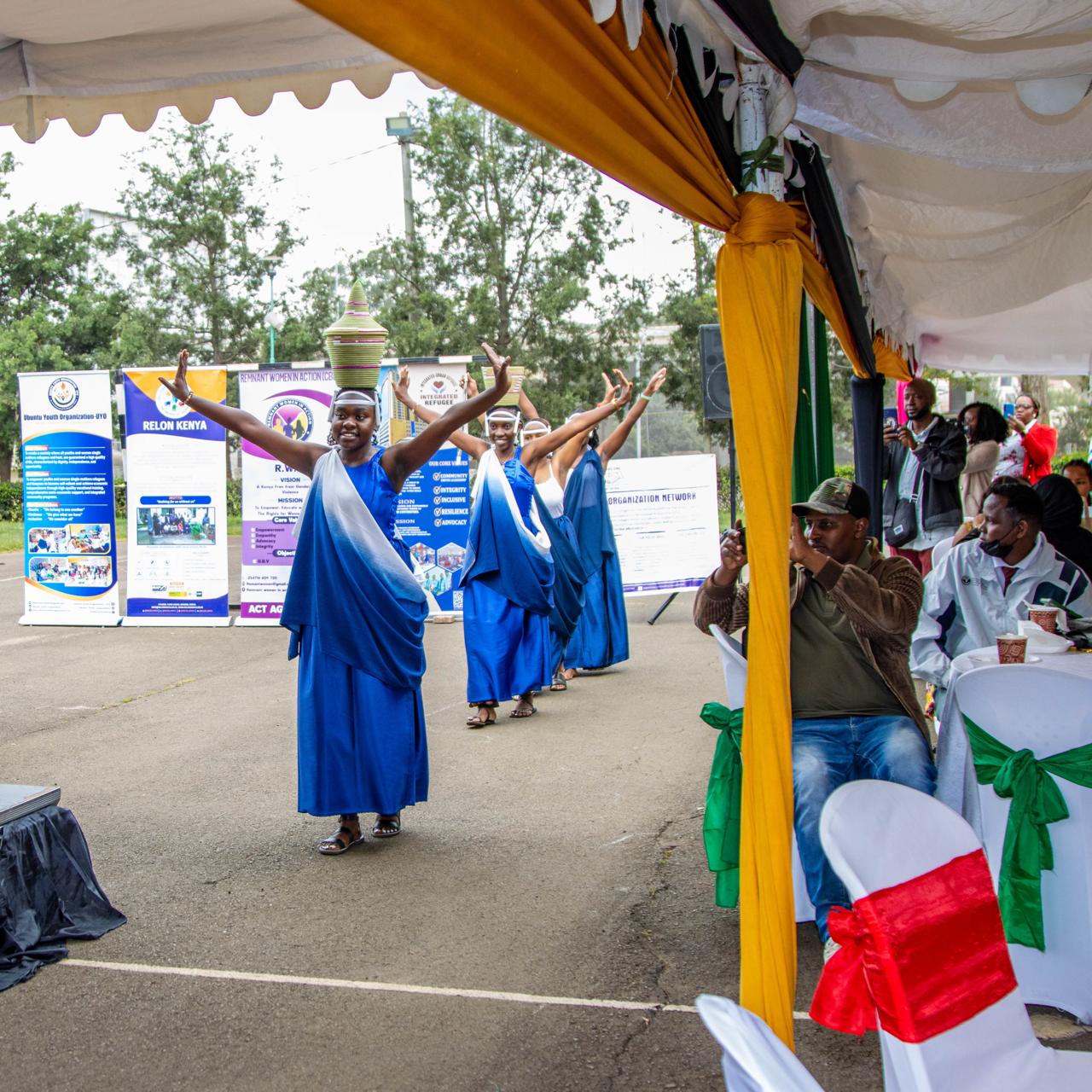 Women in coordinated blue garments performing a processional dance as guests watch beneath a decorated festival tent
